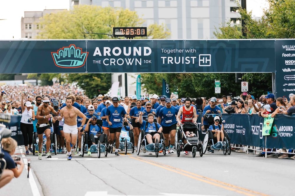 Runners and runners pushing wheelchairs crossing the start line of a road race with city buildings in the background