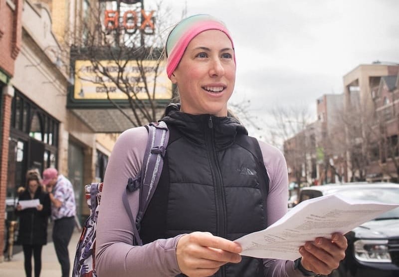 A woman outside on a city street wearing a purple long-sleeve shirt, black vest, and colorful headband and holding a paper in front of her