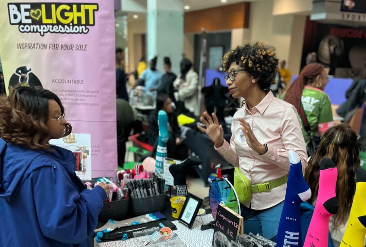A woman on one side of a table with socks displayed on it, talking to a woman on the other side of the table, inside a convention center with other people in the background