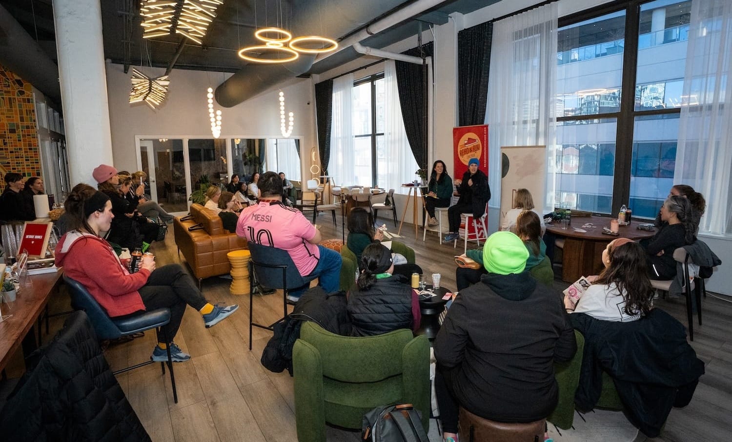 People sitting in chairs inside a room listening to two speakers at the front of the room sitting in tall chairs
