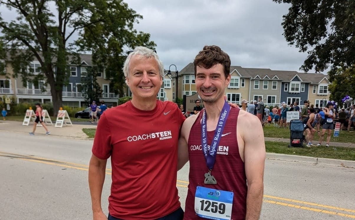 A running coach smiling and standing next to his athlete wearing a race bib and medal outside on a street on a cloudy day