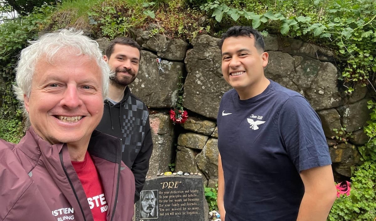 A man in a red jacket taking a selfie with two men in the background in front of a rock wall with greenery and a memorial plaque