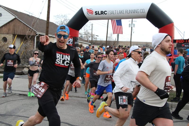 Runners starting a road race with one runner wearing black clothes and sunglasses smiling and pointing at the camera 