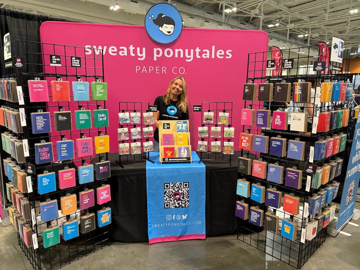 A woman standing behind a table at an expo with greeting card displays on both sides of her and a pink temporary wall behind her