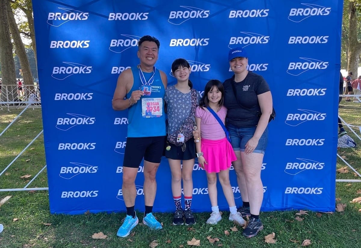 A runner holding a race medal and posing with his two daughters and wife in front of a race banner outside on the grass