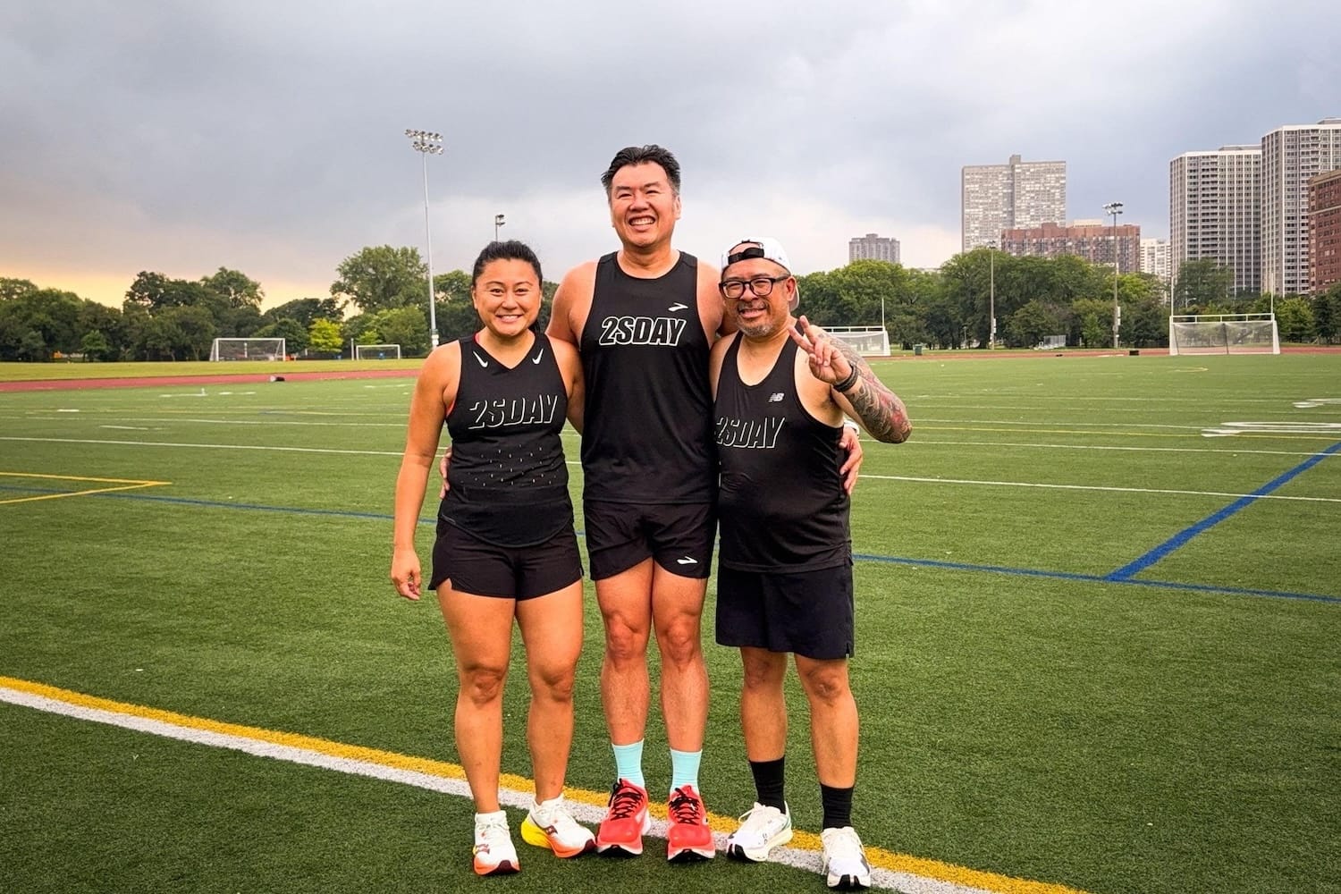 Three runners in black attire posing for a photo together on a grass field with a cloudy sky in the background