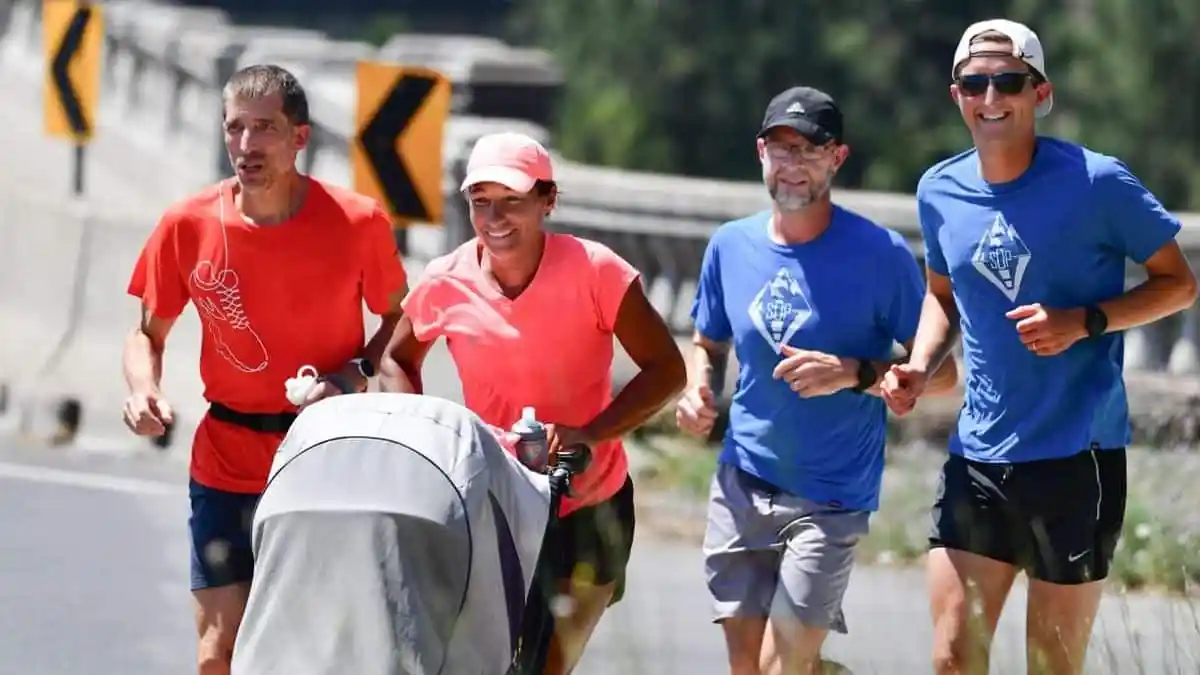 A woman in a pink shirt and hat running while pushing a gray stroller with three men running alongside her on a street