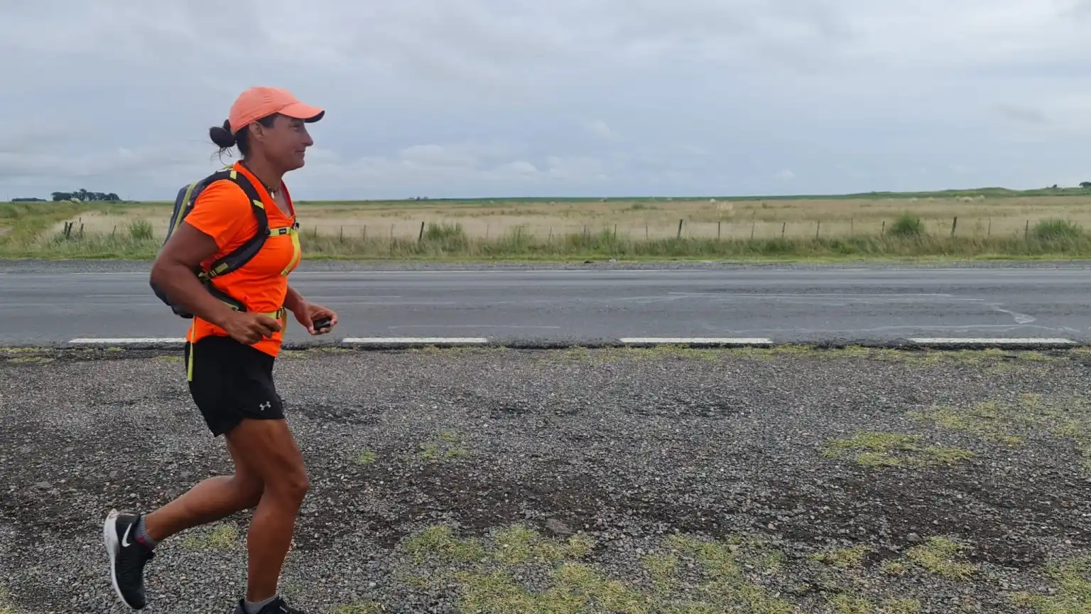 A woman in a pink shirt and hat with a small backpack running along the side of a desert road
