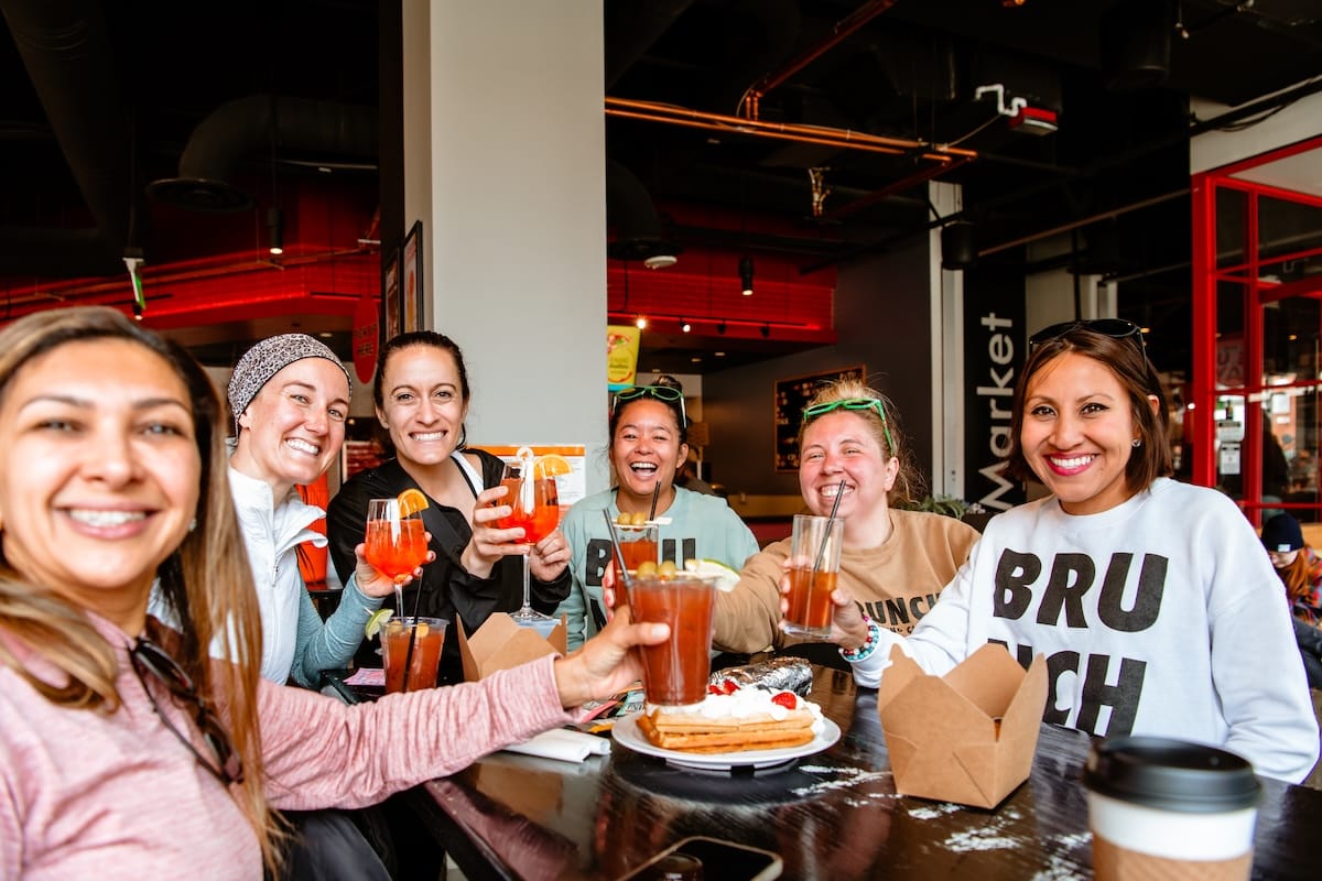 Six women each holding up a beverage glass, smiling for the camera, and sitting around a table with food dishes on it inside a restaurant