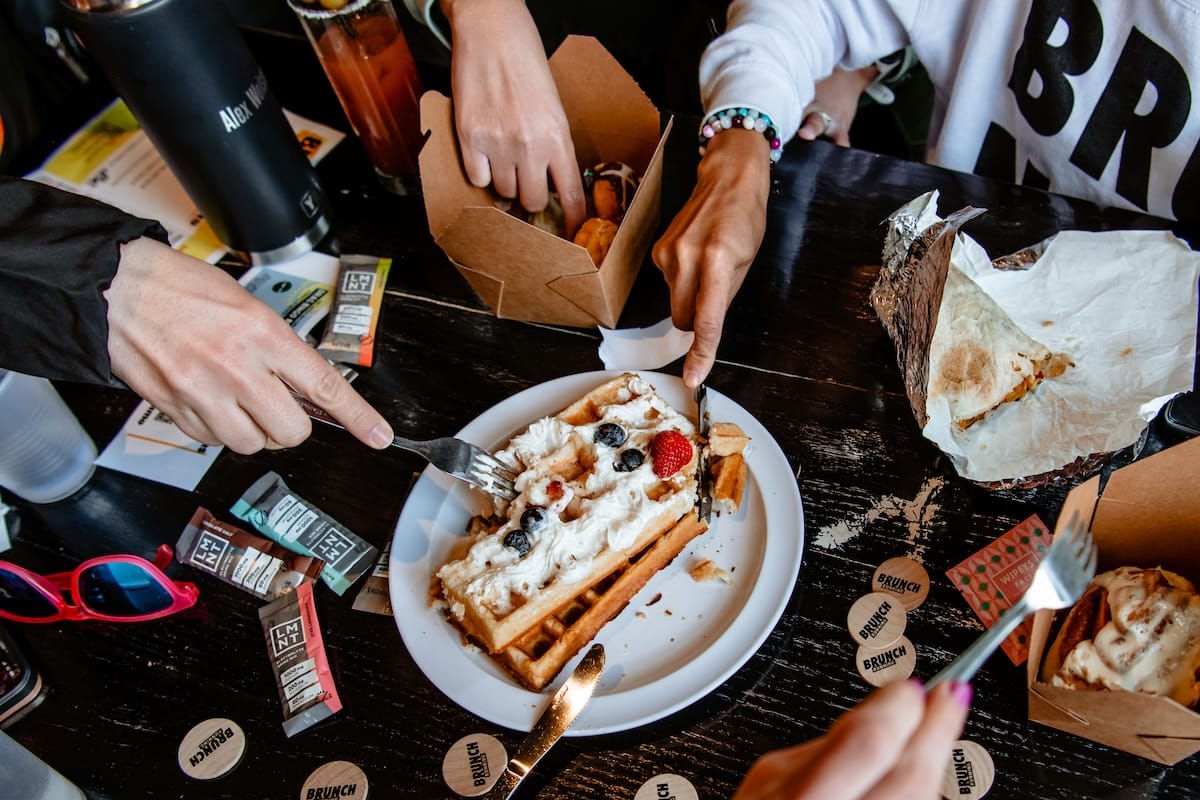 A plate of waffles topped with whipped cream and berries on a table with several hands holding forks and cutting bites of the waffle