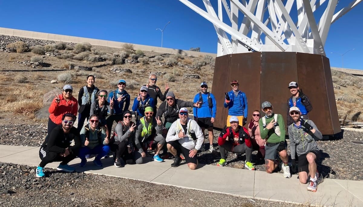 A large group of runners posing outside for a photo along a sidewalk trail with blue sky and a white art sculpture in the background