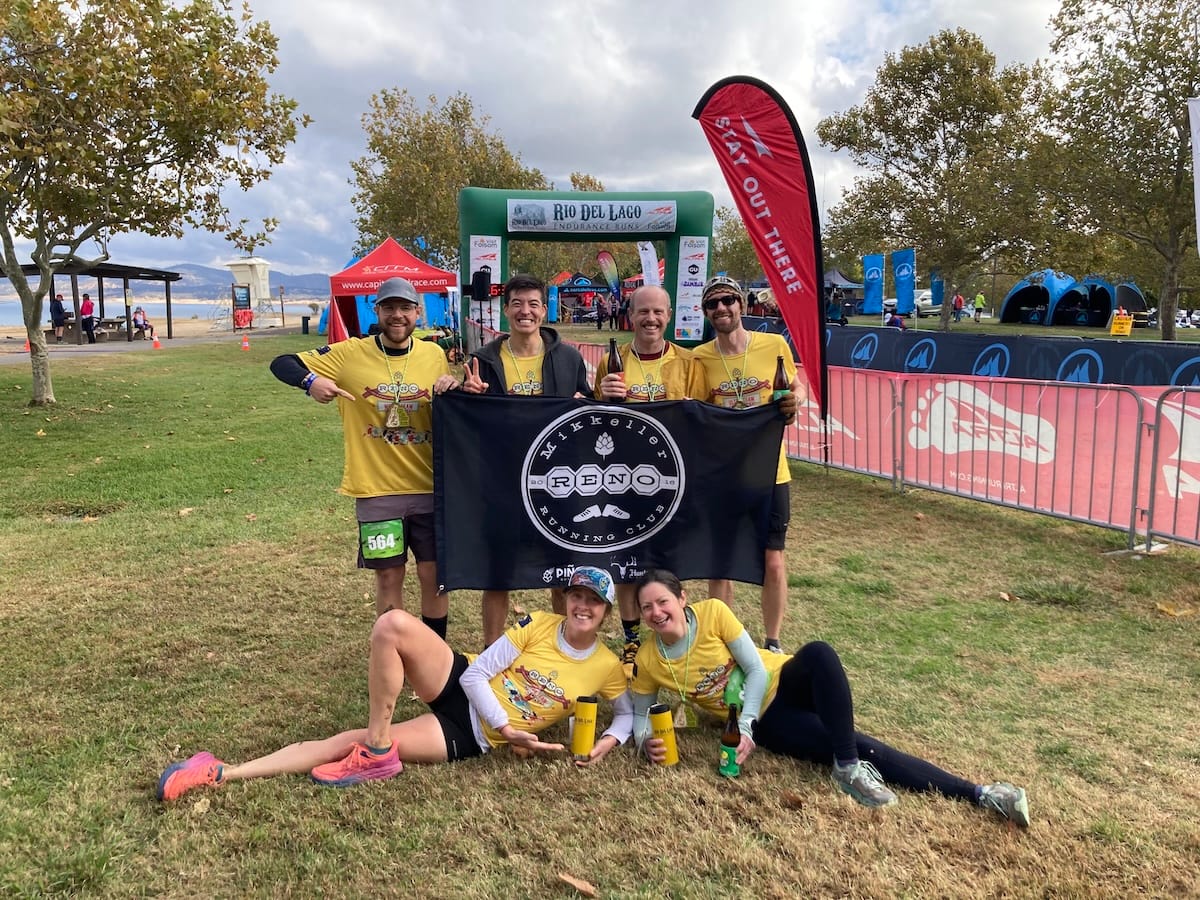 Six runners in yellow race shirts posing for a photo outside in the grass in front of a race finish line and holding a black and white banner for Mikkeller Run Club