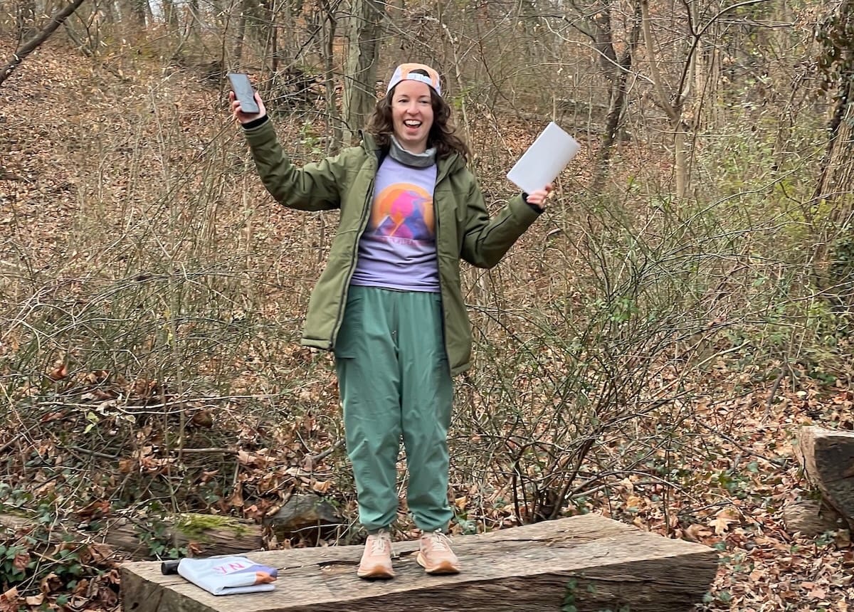 A woman standing on top of a picnic table in the forest with her arms raised and smiling
