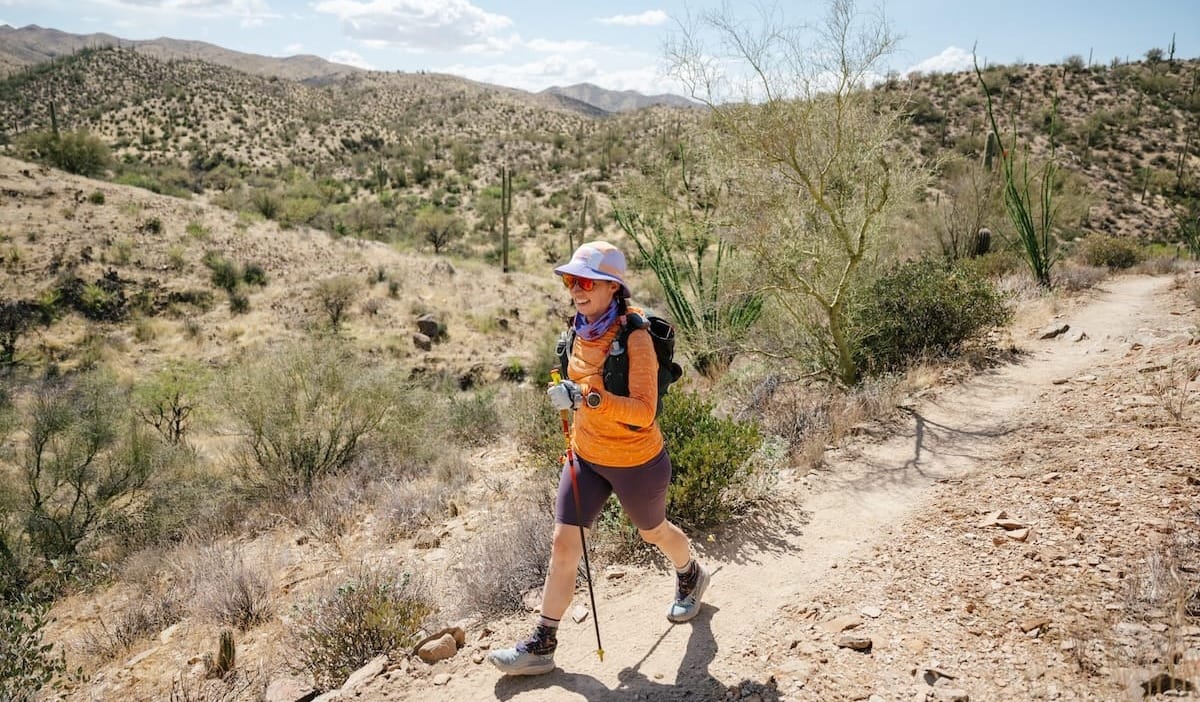 A woman hiking along a desert trail during a sunny day, wearing a hat, shorts, and long sleeves
