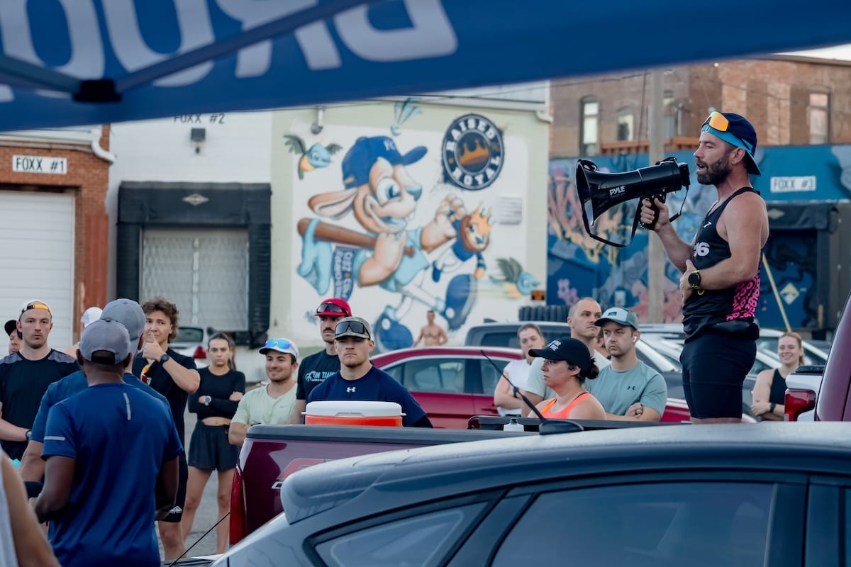 A bunch of runners standing in a parking lot in between cars with one runner speaking into a megaphone