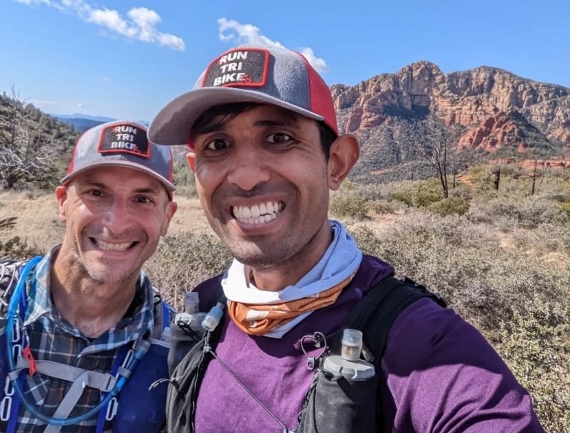 Two men wearing hats and running hydration vests posing for the camera in front of a mountain range on a sunny day