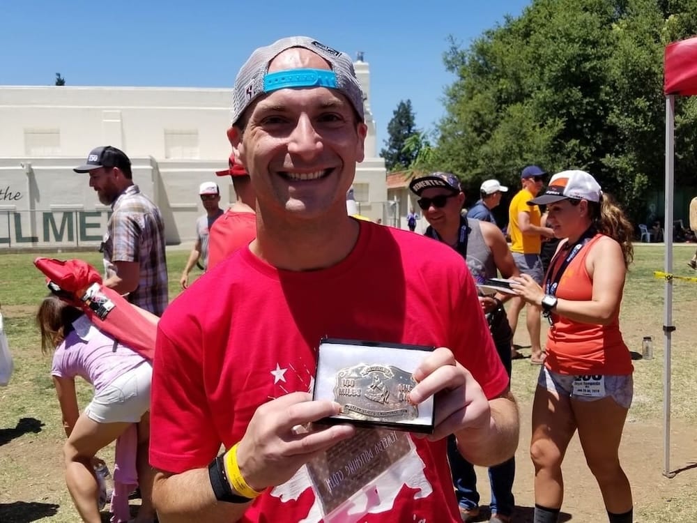 A man in a red shirt and backward ball cap holding a race finisher's award and smiling at the camera