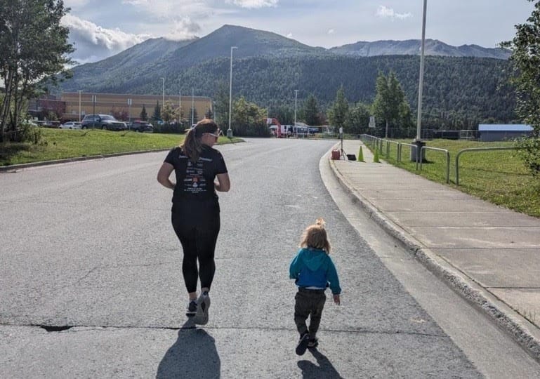 A woman and her young child running along a street away from the camera with tree-covered hills in the background