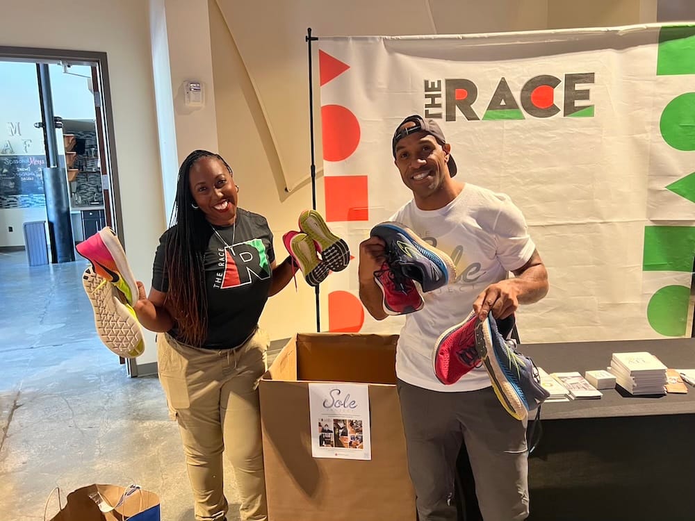 A man and a woman posing for the camera each holding a few shoes in each hand in front of a cardboard box for donations
