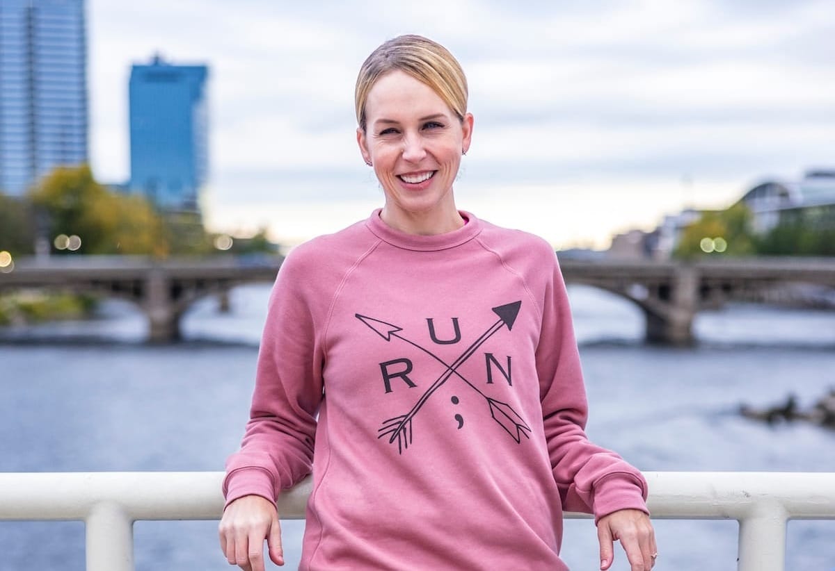 A woman in a pink sweatshirt posing for a photo on a bridge with a lake and downtown buildings in the background