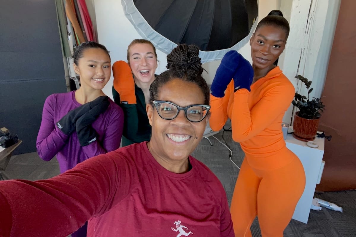 A woman taking a selfie with three women smiling behind her and showing their hands wearing mittens