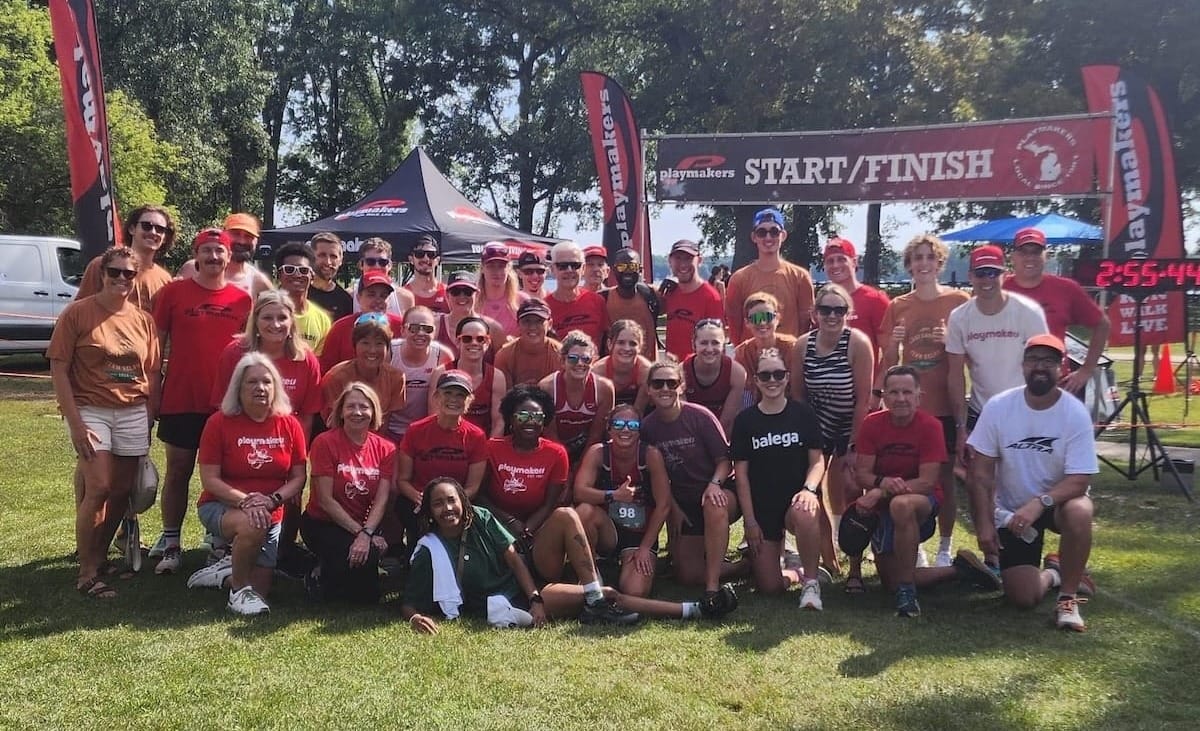 A large group of runners wearing red t-shirts gathered together for a group photo outside in the grass in front of a race start line with trees in the background on a sunny day