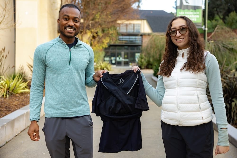 A man and a women posing together outside each holding one side of a black shirt
