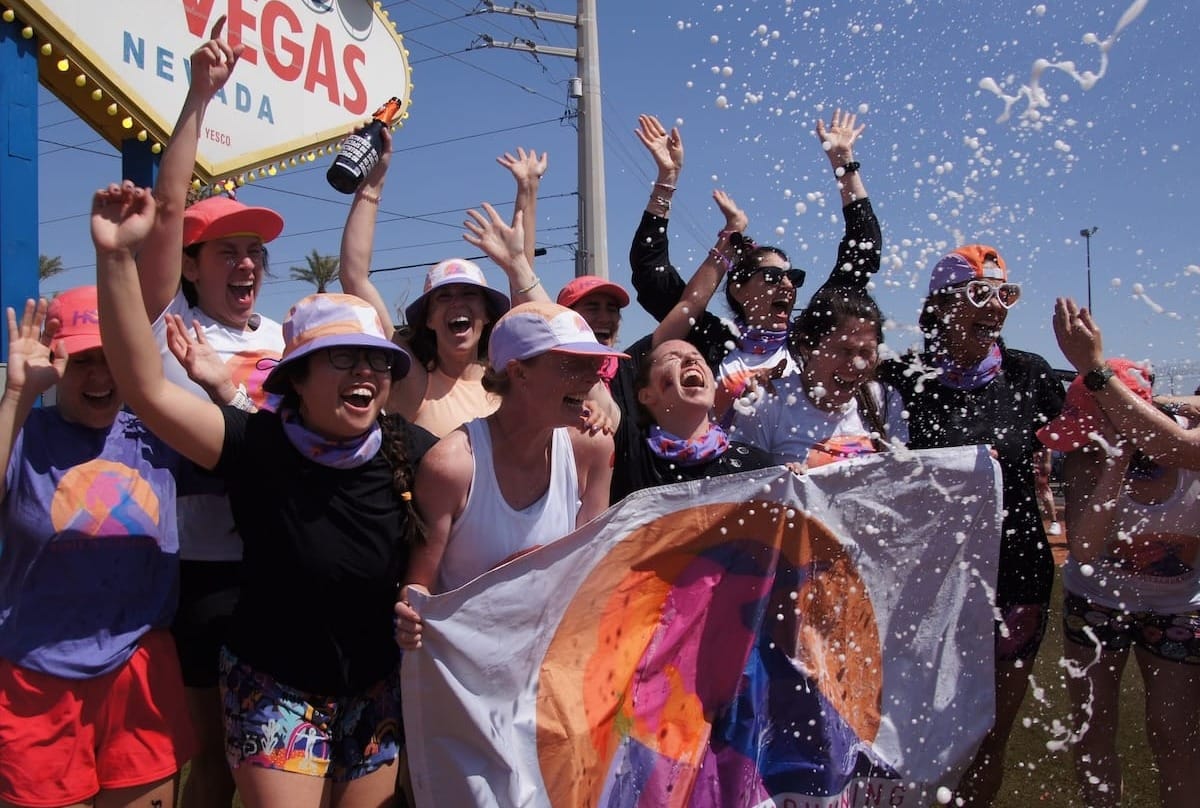 A group of women runners celebrating, holding a colorful flag in front of the Las Vegas city sign