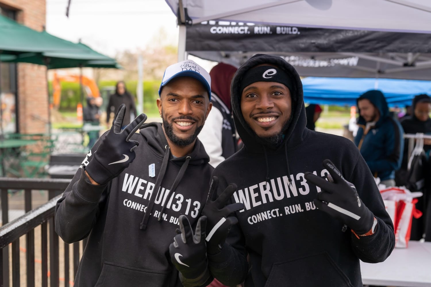 Two men in black sweatshirts holding up peace signs with their hands and posing outside in front of pop-up tents