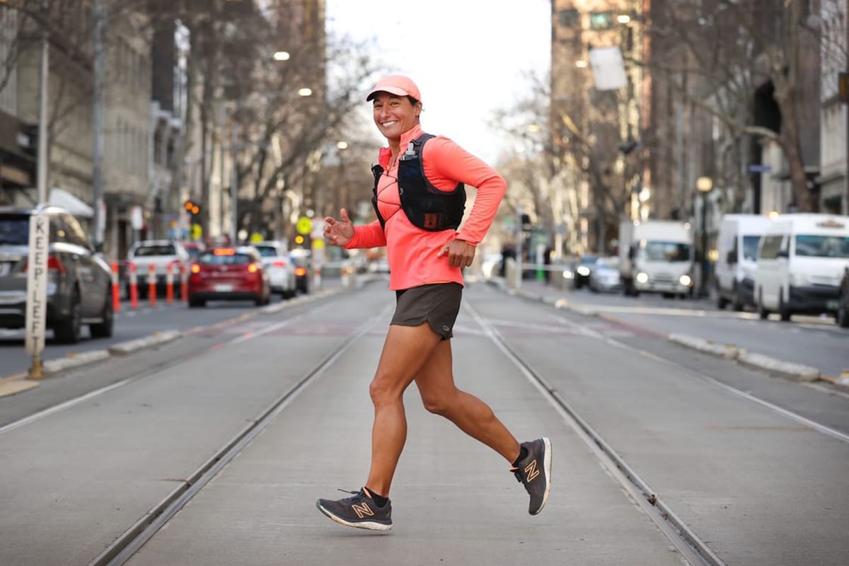 A woman in running shorts, a pink long-sleeve shirt, and a pink hat running across a downtown city street and smiling for the camera