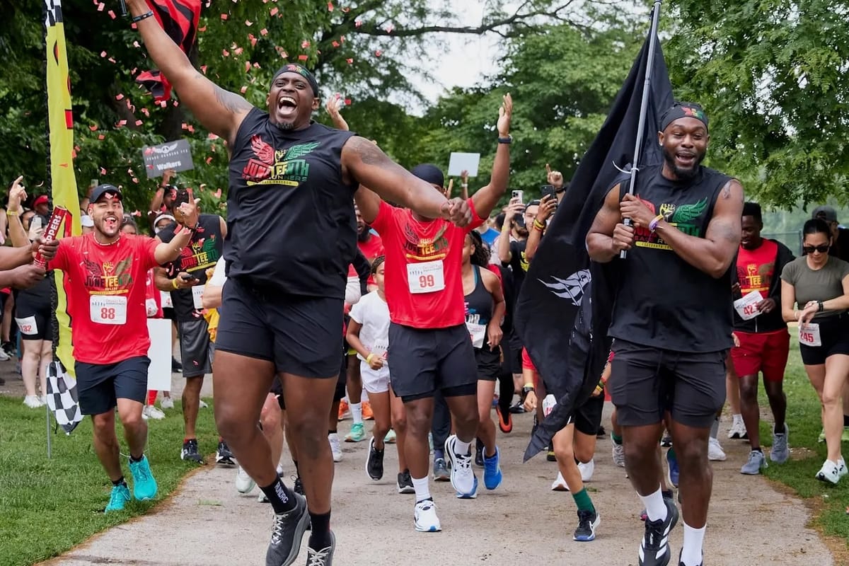 Runners wearing black and red clothing, holding flags and excitedly starting a run outside on a sidewalk with green trees in the background