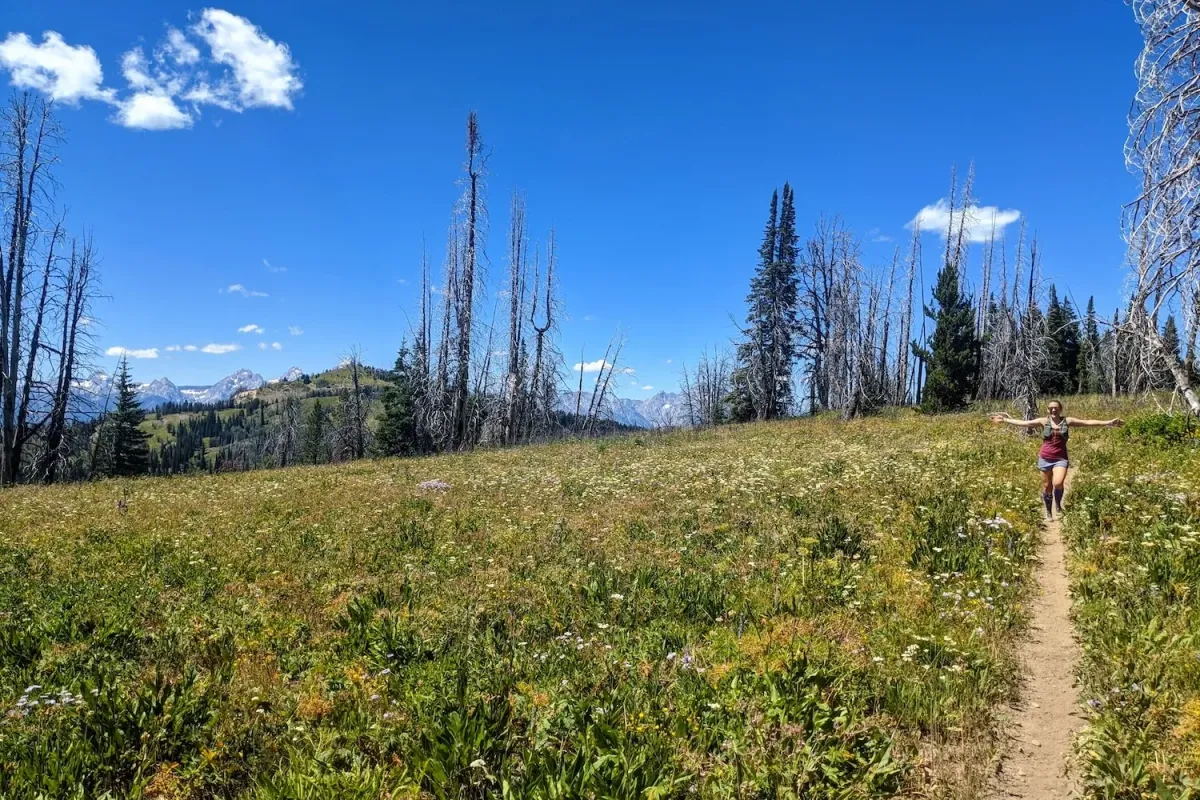 A women running along a single track trail with her arms out wide surrounded by a green meadow with a blue sky in the background