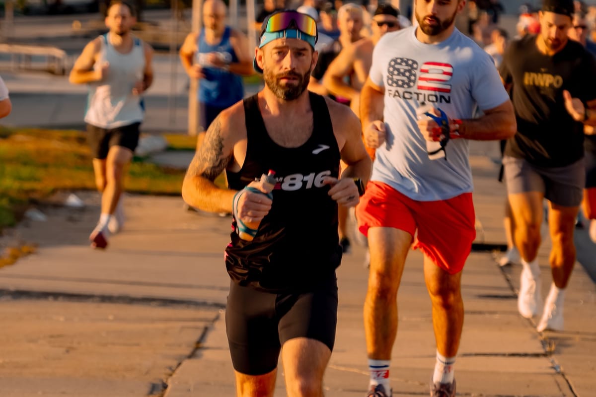 A male runner in a black tank top and shorts leading a race with other runners behind him, running on pavement