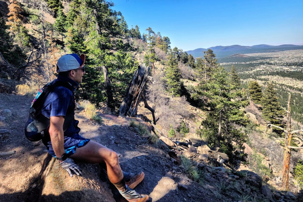 A man in activewear sitting on top of a mountain ledge looking out at the mountainous view