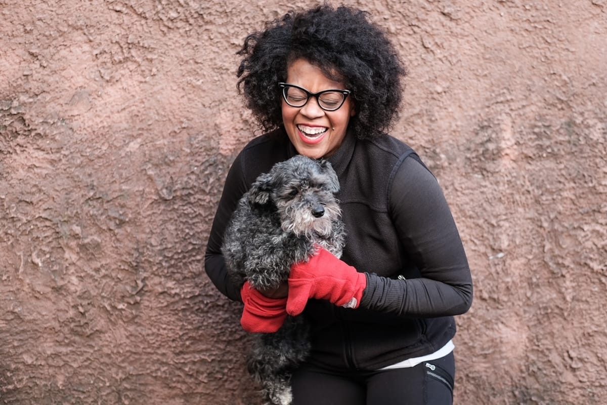 A woman laughing, wearing a black running jacket and red mittens, and holding a small dog