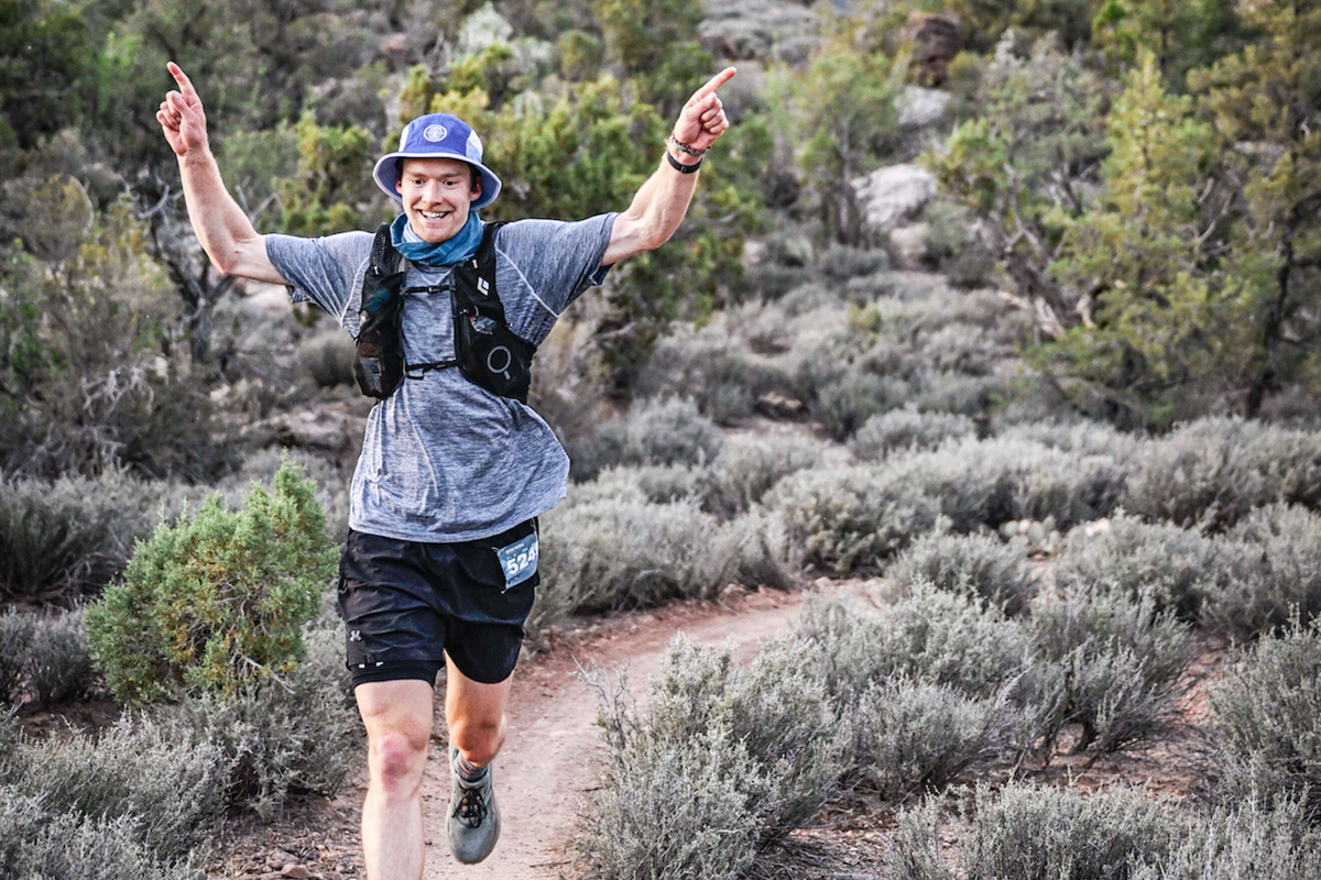 A man running along a desert trail in a shorts, a short-sleeve shirt, a hydration backpack, and a hat, smiling and raising both arms