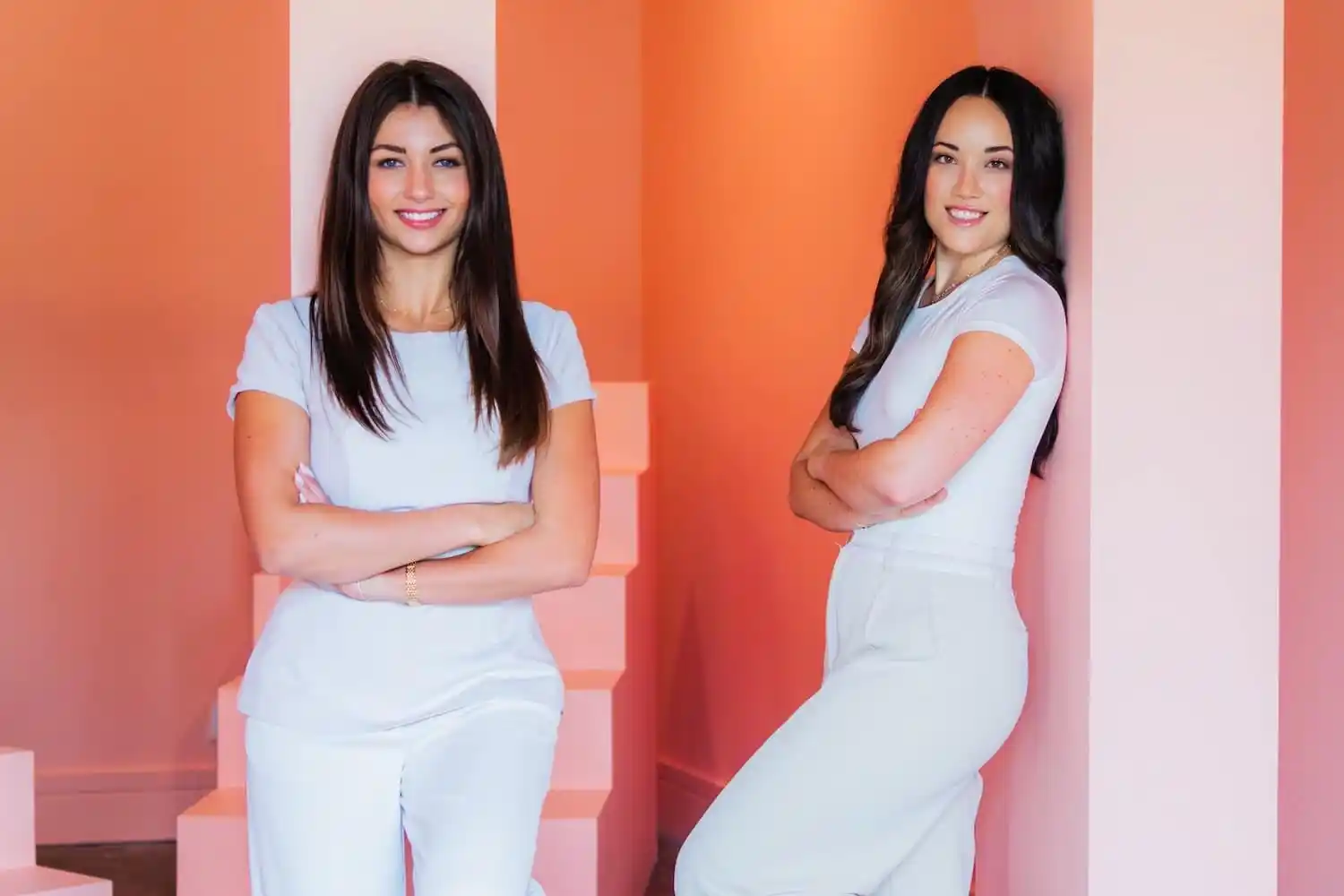 Two women in white clothes crossing their arms and posing for the camera in front of an orange and pink-hued wall