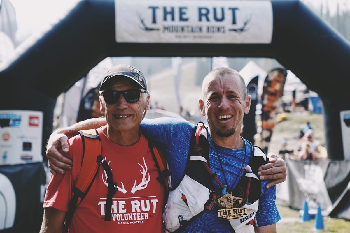 Two male runners with an arm around each other, posing for a photo in front of a race finish line