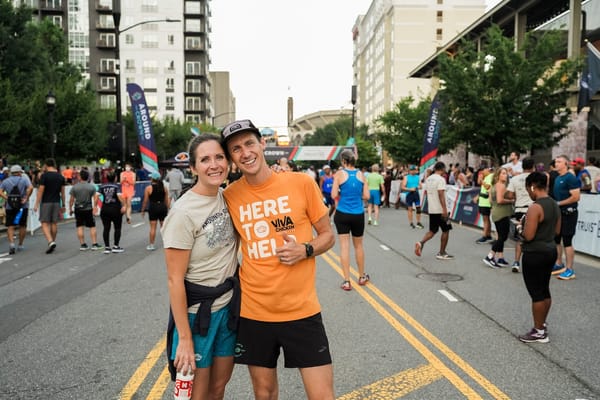 A man and a woman posing together on a street in front of a race finish line with fellow runners standing behind them