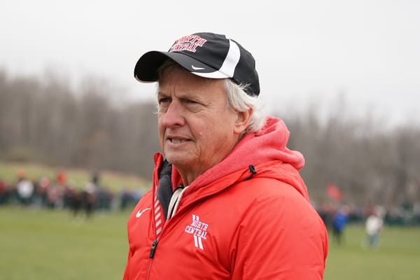 A man in a red winter jacket and a hat with a blurred background with grass, trees, and a cloudy sky