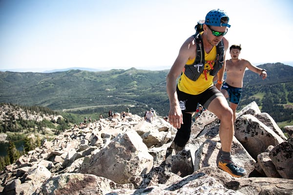 A man in a yellow tank top and a running vest hiking up a rocky mountain with others behind him and a scenic mountain background