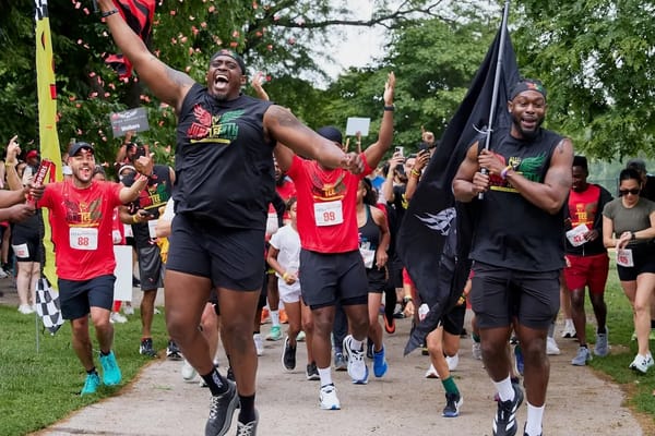 Runners wearing black and red clothing, holding flags and excitedly starting a run outside on a sidewalk with green trees in the background