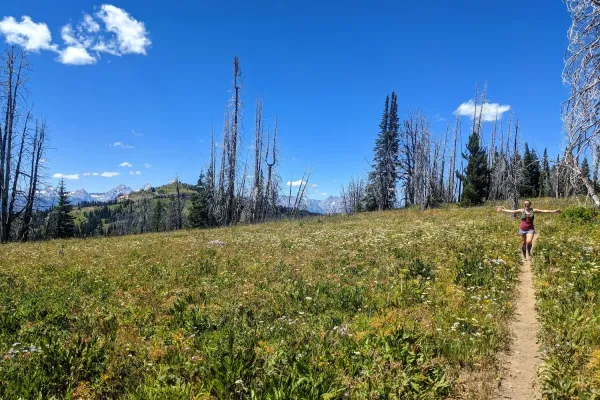 A women running along a single track trail with her arms out wide surrounded by a green meadow with a blue sky in the background