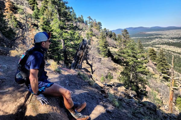 A man in activewear sitting on top of a mountain ledge looking out at the mountainous view