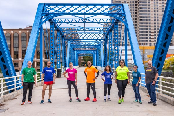 Eight people in brightly colored clothes, posing outside in a city by standing on a walkway with a blue bridge rails above them