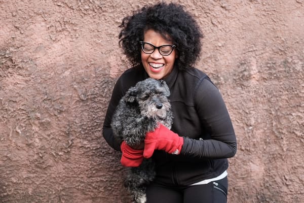 A woman laughing, wearing a black running jacket and red mittens, and holding a small dog