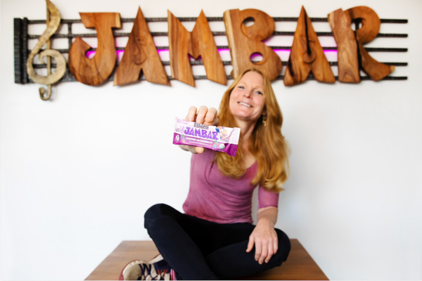 A woman sitting cross-legged on a table, smiling and holding an energy bar in a wrapper toward the camera