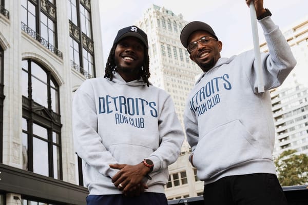 Two men in white sweatshirts and black hats, posing for a photo in front of city buildings